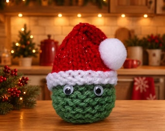 May include: A round, green, hand-knitted creature with two plastic googly eyes, wearing a red knitted hat with a white brim and a white pom-pom on top. The object is crafted from thick yarn and features a textured, chunky knit pattern.