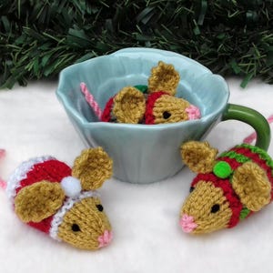 Könnte beinhalten: Handgestrickte Weihnachtsmäuse in einer hellblauen Tasse. Die Mäuse sind beige, rot und grün mit rosa Schwänzen und Ohren. Eine Maus trägt einen Weihnachtsmütze. Die Tasse steht auf einer weißen Oberfläche mit grüner Girlande im Hintergrund.