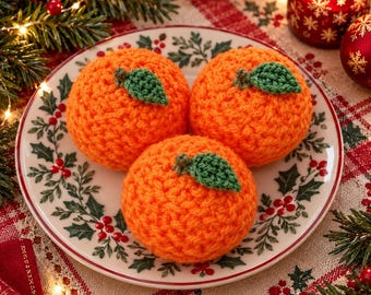 May include: Three round, bright orange crocheted spheres, each topped with a small, textured green leaf. These handmade yarn fruits are arranged on a white ceramic plate decorated with a pattern of green holly leaves and red berries.