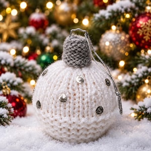 May include: A spherical Christmas ornament covered in a white knitted fabric with a textured grey knitted cap at the top. The white surface is decorated with several small, round, metallic silver studs. A thin silver string is attached for hanging.