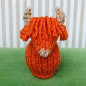 May include: A small, orange knitted toy cow with brown ears. The cow is sitting on a green surface.