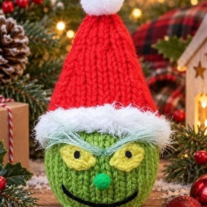 May include: A round, green knitted character ornament featuring a red knitted Santa hat with a white pom-pom and a fuzzy white trim. The character has yellow knitted eyes with black pupils, a small green pom-pom nose, and a stitched black smile.