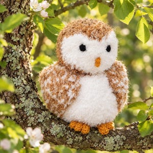 May include: A crocheted owl plush toy featuring a round body with textured brown and white yarn, two black circular eyes, a small orange beak, and two orange feet, perched on a tree branch with green leaves and white blossoms.
