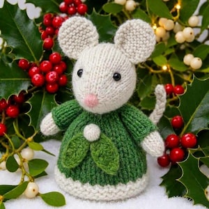 Op de afbeelding: Een handgebreid muisfiguurtje van crèmekleurig garen met ronde oren en een klein roze neusje. De muis draagt een groen gebreid jurkje met een witte pompon en twee groene bladvormige accenten aan de voorzijde. De muis heeft een lange, gevlochten staart.