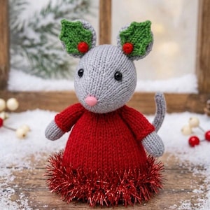 Peut inclure: Une figurine de souris tricotée grise portant un pull rouge avec une bordure en guirlande rouge métallique à la base. La souris a de grandes oreilles décorées de feuilles de houx tricotées vertes et de petites baies rouges, des yeux en perles noires et un nez rose.