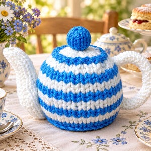 Op de afbeelding: Een ronde, gehaakte theemuts met horizontale strepen van felblauw en wit garen. De theemuts heeft een effen blauwe, bolvormige knop, een wit handvat en een witte tuitafdekking, ontworpen om over een standaard theepot te passen.
