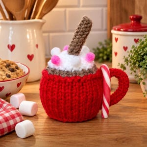 May include: A crocheted mug cosy in bright red featuring a white yarn topping with small pink pom-poms, a brown crocheted stirrer, and a red and white striped candy cane attached to the side handle, creating a festive hot cocoa appearance.