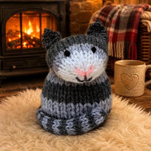 May include: A hand-knitted cat figurine featuring a striped pattern in shades of dark gray, light gray, and white. The cat has two pointed ears, small black bead eyes, a stitched pink nose, and a curled tail resting at the base of its body.