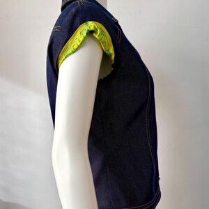May include: Dark blue denim vest with a collared neckline and short sleeves. The sleeve cuffs are lined with a bright yellow fabric featuring a green geometric pattern. The vest has visible yellow stitching.