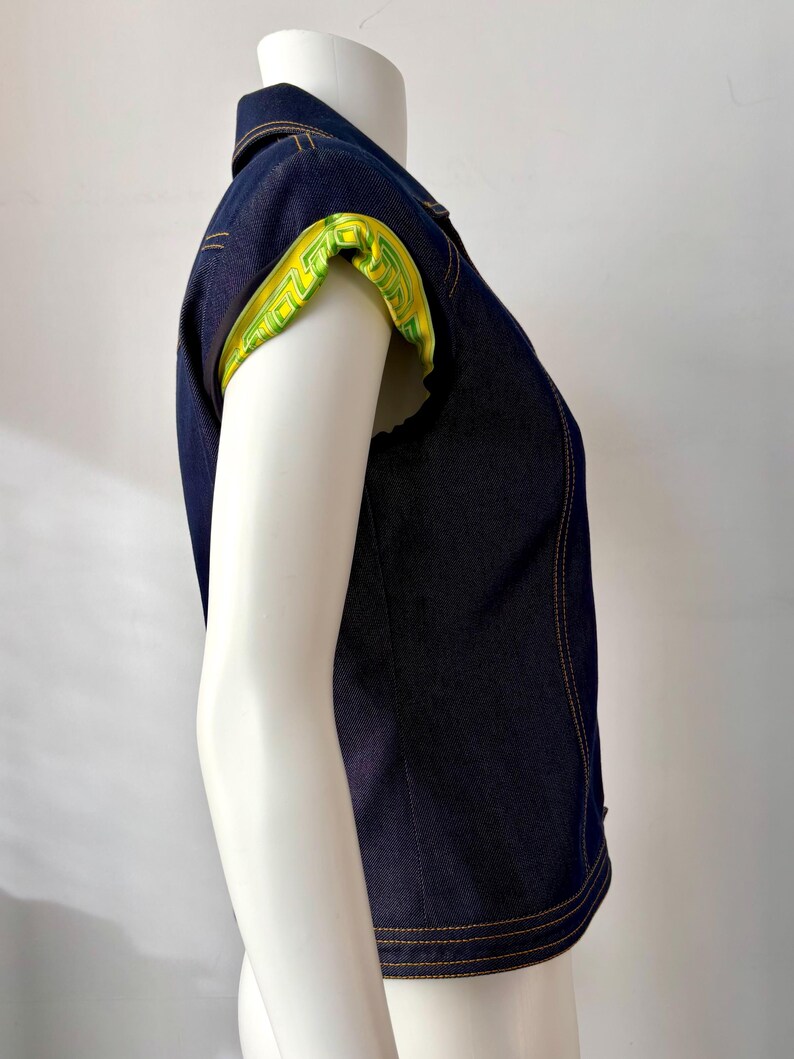 May include: Dark blue denim vest with a collared neckline and short sleeves. The sleeve cuffs are lined with a bright yellow fabric featuring a green geometric pattern. The vest has visible yellow stitching.