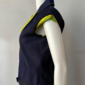 May include: A sleeveless denim vest with a collared neckline. The vest features yellow and green patterned fabric on the inner collar and sleeve openings. The vest is dark blue with visible stitching.