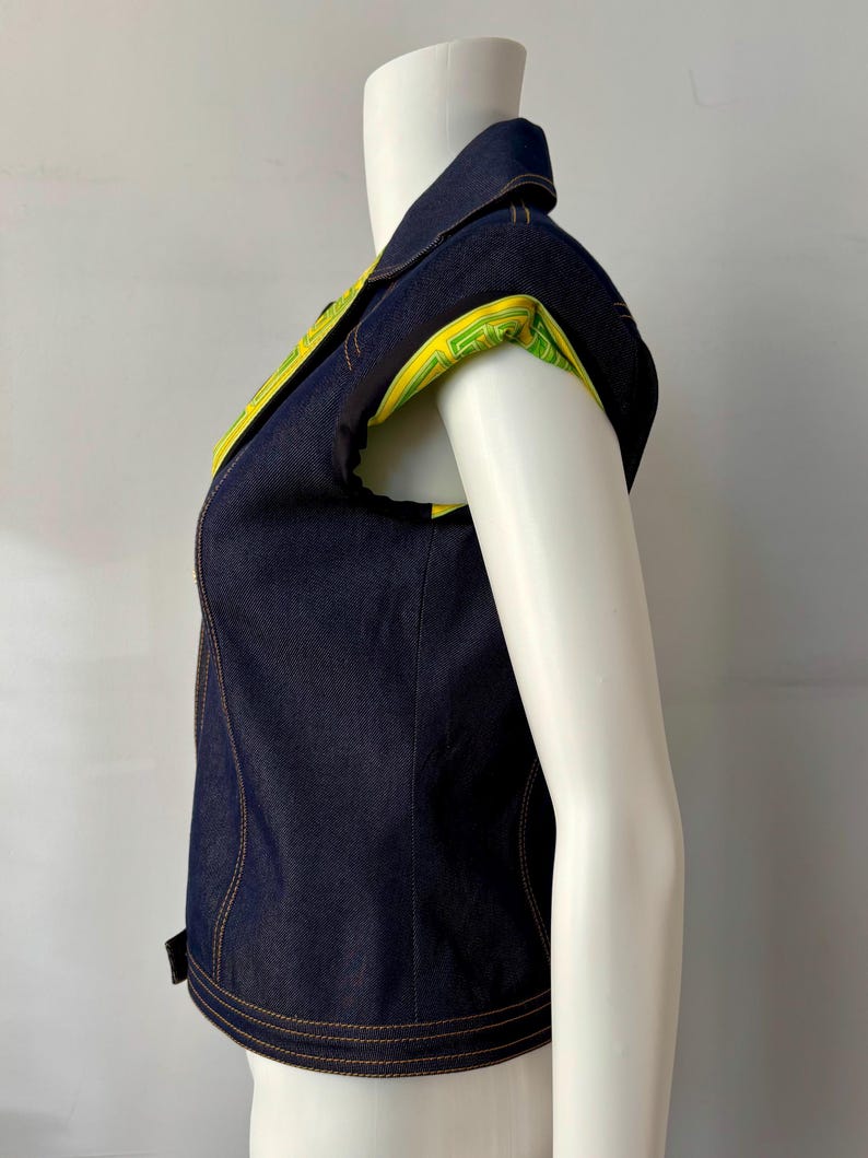 May include: A sleeveless denim vest with a collared neckline. The vest features yellow and green patterned fabric on the inner collar and sleeve openings. The vest is dark blue with visible stitching.