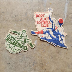 Op de afbeelding: Twee vintage softbalpatches. Eén patch heeft een groen en wit ontwerp met een softbalspeler en de tekst "PORT ALICE SOFTBALL 1948". De andere patch toont een speler in blauw en rood met de tekst "PORT ALICE SOFTBALL CLUB".