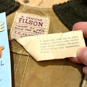 Pu&ograve; includere: Primo piano di una giacca Filson color cachi con colletto verde scuro. La giacca presenta un'etichetta cucita con la scritta "A Genuine Filson" e istruzioni per la cura. Il tessuto della giacca &egrave; visibile.
