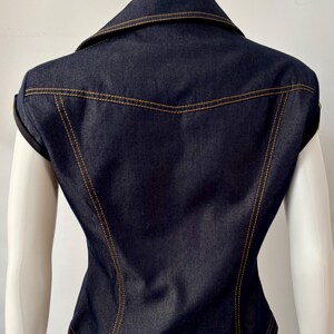May include: Dark blue denim vest with a collared neckline and gold stitching. The vest has a fitted silhouette and is sleeveless. The back of the vest is visible, showcasing the tailored design and stitching details.