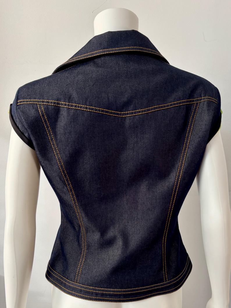 May include: Dark blue denim vest with a collared neckline and gold stitching. The vest has a fitted silhouette and is sleeveless. The back of the vest is visible, showcasing the tailored design and stitching details.