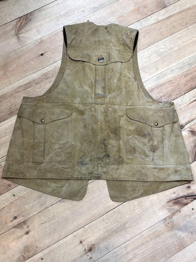 Pu&ograve; includere: Un gilet color cachi con pi&ugrave; tasche e chiusura con bottoni. Il gilet ha un aspetto vissuto, che suggerisce che sia realizzato in un materiale resistente. Presenta due ampie tasche sul davanti e una tasca pi&ugrave; piccola sul petto.