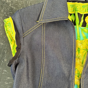 May include: A dark blue denim vest with yellow stitching. The vest has a collar and a colorful patterned lining with the "Gianni Versace" label visible. The lining features a green and yellow floral design.