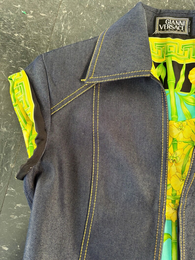 May include: A dark blue denim vest with yellow stitching. The vest has a collar and a colorful patterned lining with the "Gianni Versace" label visible. The lining features a green and yellow floral design.