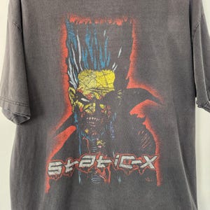 Puede incluir: Camiseta gris oscuro con un gráfico de una figura similar a un zombi con detalles en amarillo, azul y rojo. El texto "STATIC-X" está impreso en rojo. La camiseta tiene un aspecto vintage y desgastado.