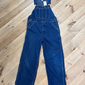 May include: Blue denim overalls with adjustable straps, a bib pocket, and multiple pockets. The workwear-style garment features metal hardware and a classic design. The overalls are displayed on a wooden surface.