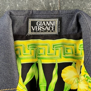 May include: Close-up of a denim jacket collar with a vibrant patterned lining. The lining features a black background with green bamboo stalks, yellow flowers, and geometric designs. The label reads "GIANNI VERSACE COUTURE MADE IN ITALY."