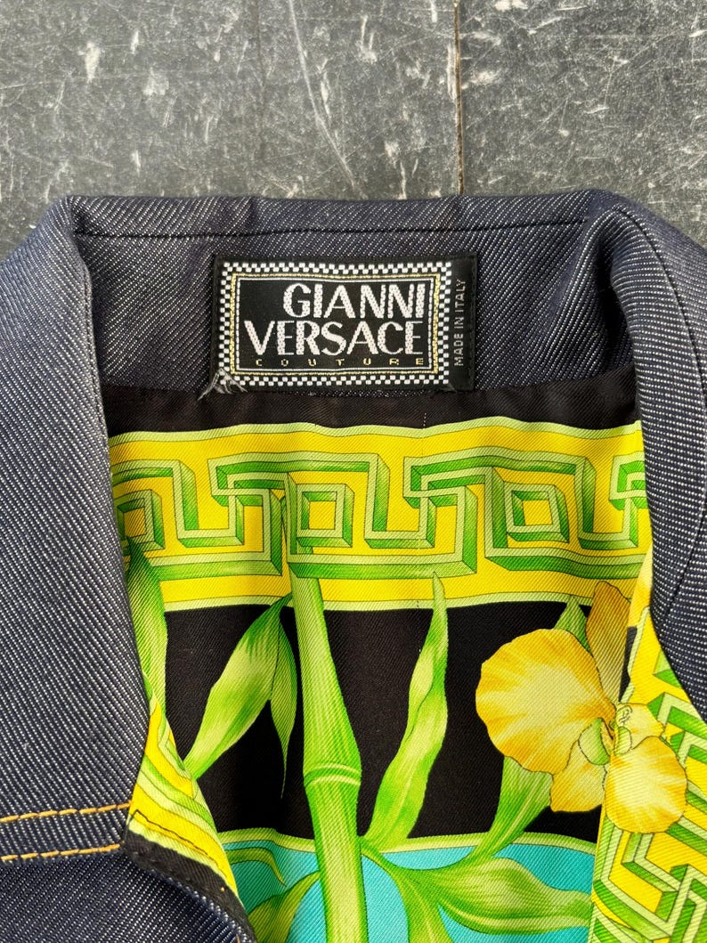 May include: Close-up of a denim jacket collar with a vibrant patterned lining. The lining features a black background with green bamboo stalks, yellow flowers, and geometric designs. The label reads "GIANNI VERSACE COUTURE MADE IN ITALY."