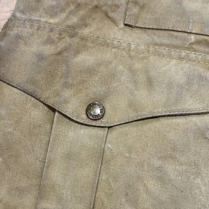 Pu&ograve; includere: Primo piano di un capo color beige, possibilmente una giacca o una camicia, con cuciture visibili e una tasca. Un bottone di metallo &egrave; centrato sulla patta della tasca. Il tessuto ha un aspetto usurato e strutturato.