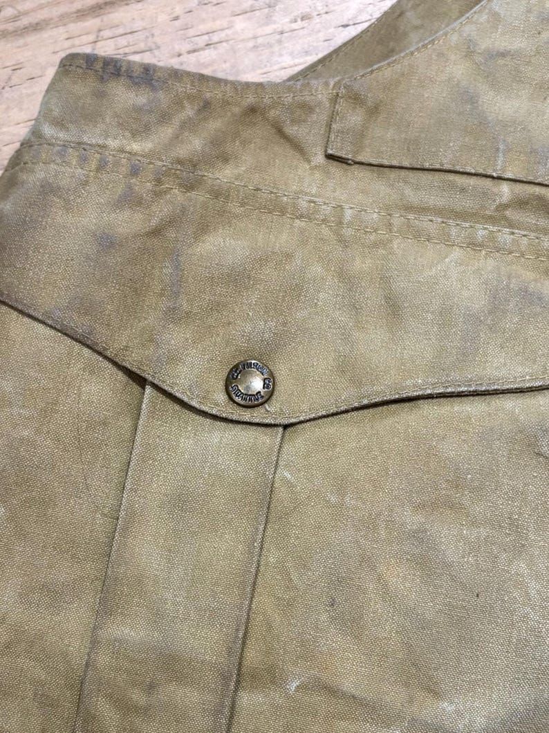 Pu&ograve; includere: Primo piano di un capo color beige, possibilmente una giacca o una camicia, con cuciture visibili e una tasca. Un bottone di metallo &egrave; centrato sulla patta della tasca. Il tessuto ha un aspetto usurato e strutturato.