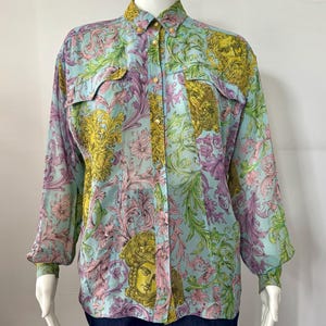 May include: A long-sleeved button-down shirt with a colorful floral and Medusa head pattern. The shirt features a light blue background with pink, yellow, and green accents. It has two chest pockets and gold-colored buttons.