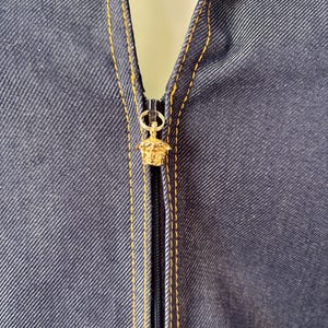 May include: Close-up of a dark blue denim garment with a gold-colored zipper. The zipper pull features a gold-colored Medusa head design. The garment has gold stitching along the seams.