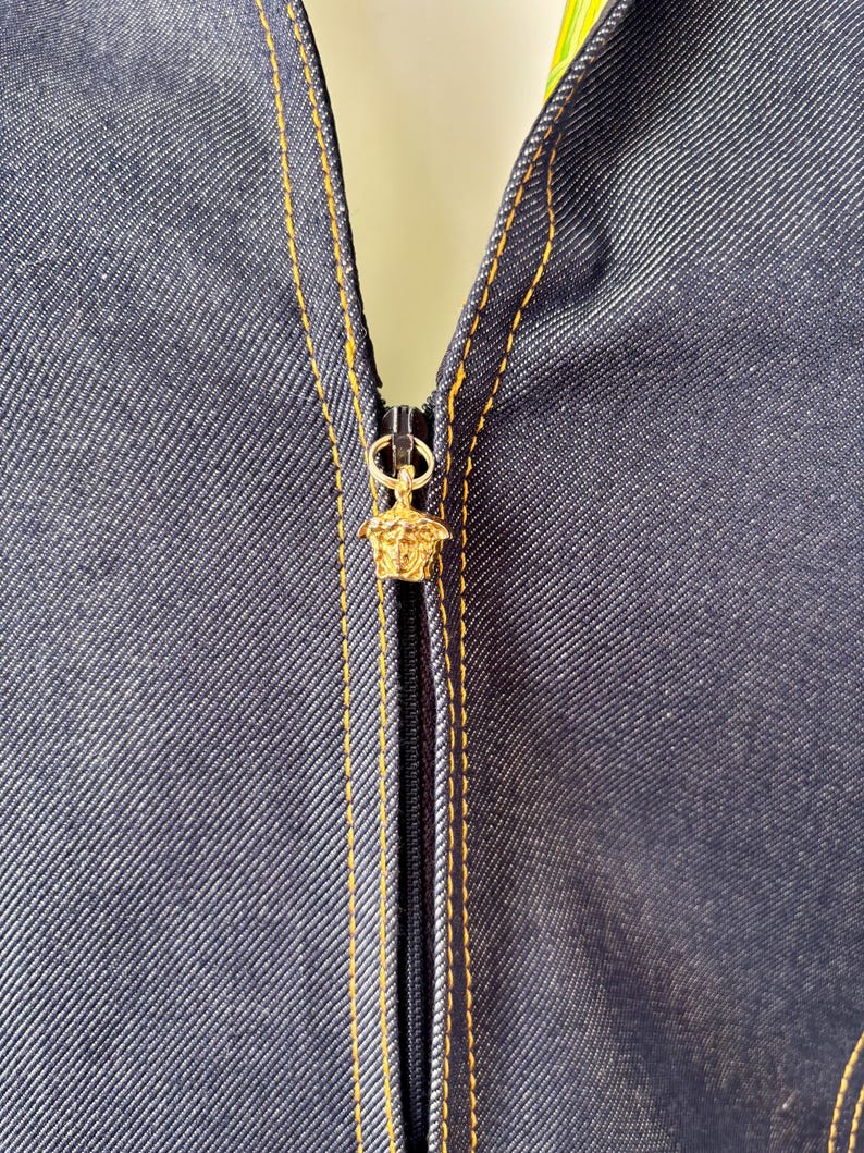 May include: Close-up of a dark blue denim garment with a gold-colored zipper. The zipper pull features a gold-colored Medusa head design. The garment has gold stitching along the seams.
