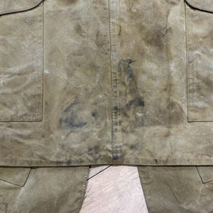 Pu&ograve; includere: Primo piano di un grembiule usurato, color beige, con due ampie tasche e un lembo inferiore. Il tessuto mostra segni di usura e macchie, con macchie pi&ugrave; scure e una cucitura verticale al centro. Il grembiule &egrave; probabilmente per lavoro o utilit&agrave;.