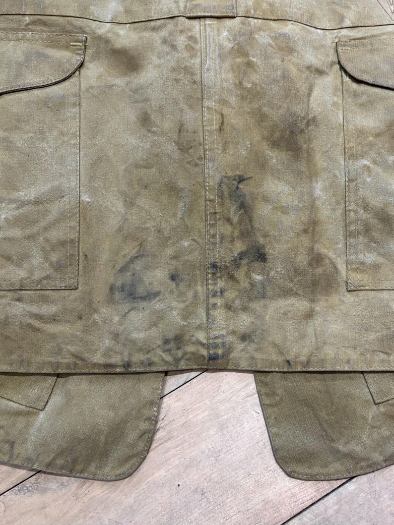 Pu&ograve; includere: Primo piano di un grembiule usurato, color beige, con due ampie tasche e un lembo inferiore. Il tessuto mostra segni di usura e macchie, con macchie pi&ugrave; scure e una cucitura verticale al centro. Il grembiule &egrave; probabilmente per lavoro o utilit&agrave;.