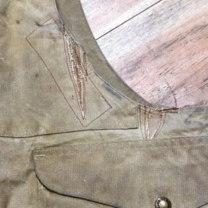 Pu&ograve; includere: Primo piano di un gilet beige con una tasca e un bottone. Il gilet mostra segni di usura, con cuciture e scolorimento visibili. Il bottone &egrave; di colore oro con un design circolare.