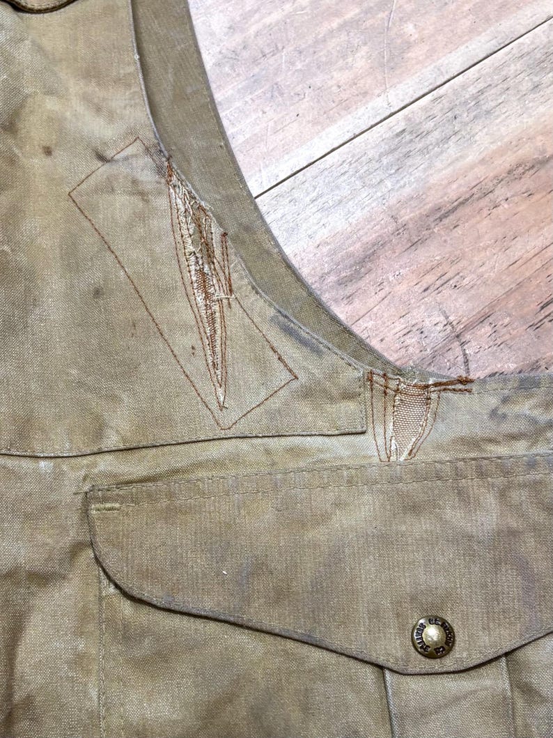 Pu&ograve; includere: Primo piano di un gilet beige con una tasca e un bottone. Il gilet mostra segni di usura, con cuciture e scolorimento visibili. Il bottone &egrave; di colore oro con un design circolare.