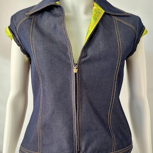 May include: A dark blue denim zip-up top with a collar and short sleeves. The top features gold stitching, a gold zipper pull, and a decorative button at the hem. The inside of the collar and sleeves are lined with a yellow and green patterned fabric.