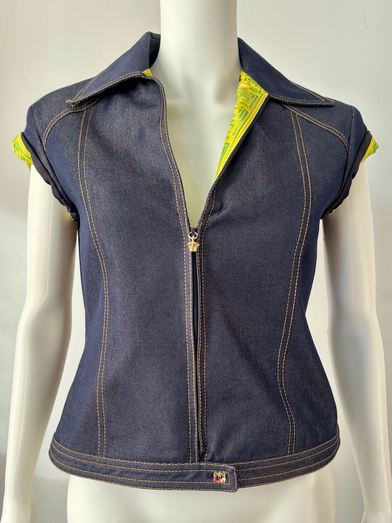 May include: A dark blue denim zip-up top with a collar and short sleeves. The top features gold stitching, a gold zipper pull, and a decorative button at the hem. The inside of the collar and sleeves are lined with a yellow and green patterned fabric.