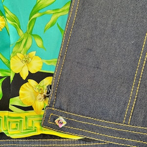 May include: Close-up of a denim jacket with yellow stitching and a colorful floral silk lining. The lining features yellow orchids and green bamboo against a teal background. A decorative button with pink and purple accents is visible.