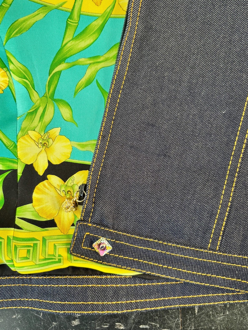 May include: Close-up of a denim jacket with yellow stitching and a colorful floral silk lining. The lining features yellow orchids and green bamboo against a teal background. A decorative button with pink and purple accents is visible.