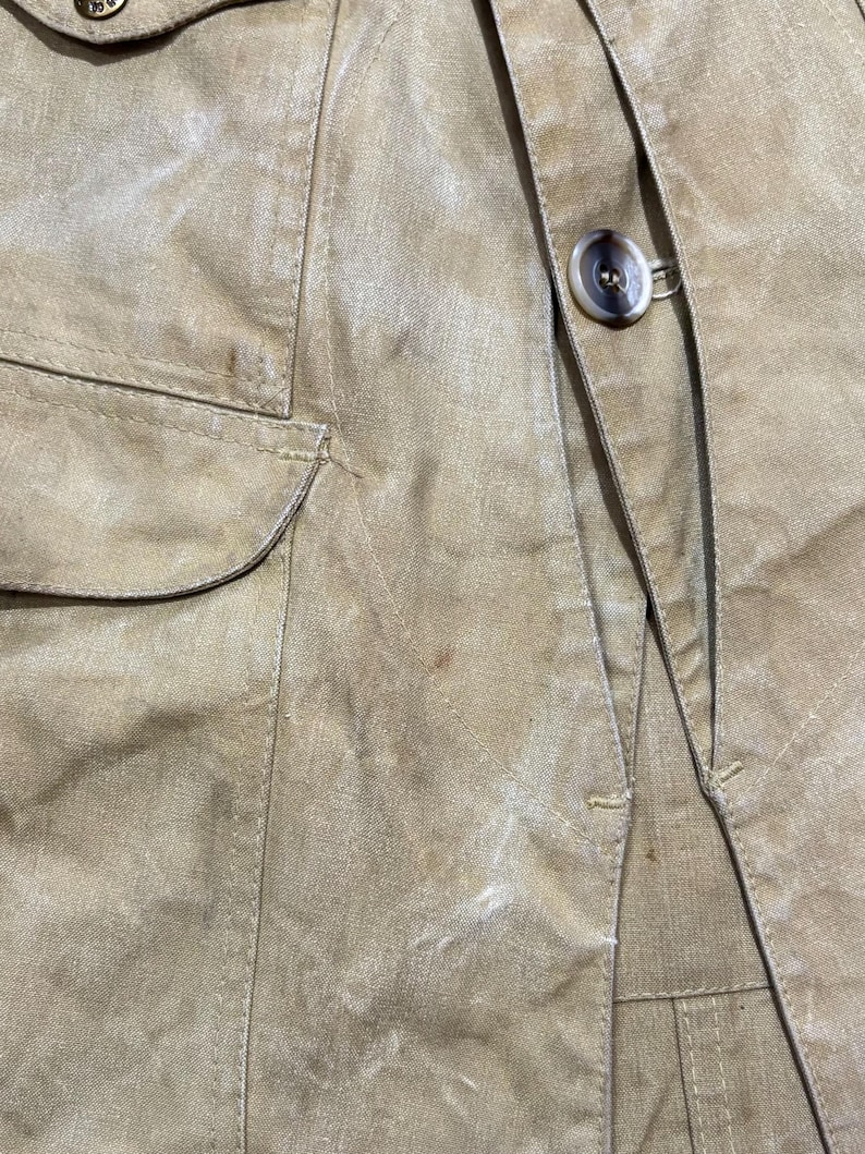 Pu&ograve; includere: Primo piano di una giacca beige con chiusura a bottoni e una tasca. La giacca ha un aspetto strutturato e dettagli di cucitura visibili. Il bottone &egrave; marrone chiaro.