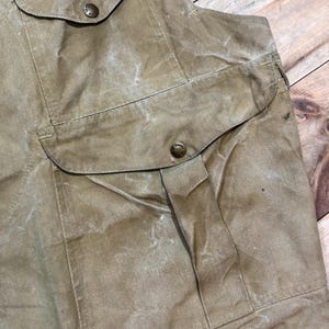 Pu&ograve; includere: Primo piano di un gilet beige con due grandi tasche frontali e due tasche sul petto pi&ugrave; piccole. Le tasche sono fissate con bottoni rotondi in metallo. Il gilet ha un aspetto usurato e strutturato.