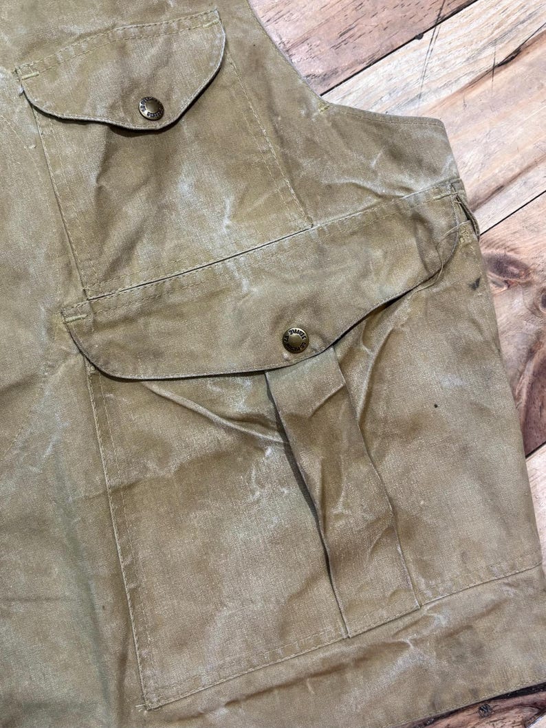 Pu&ograve; includere: Primo piano di un gilet beige con due grandi tasche frontali e due tasche sul petto pi&ugrave; piccole. Le tasche sono fissate con bottoni rotondi in metallo. Il gilet ha un aspetto usurato e strutturato.