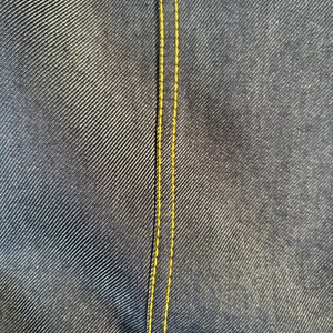 May include: Close-up of dark blue denim fabric with a yellow stitched seam running vertically. The denim has a textured appearance, with diagonal lines creating a subtle pattern. The yellow stitching provides a contrasting accent.
