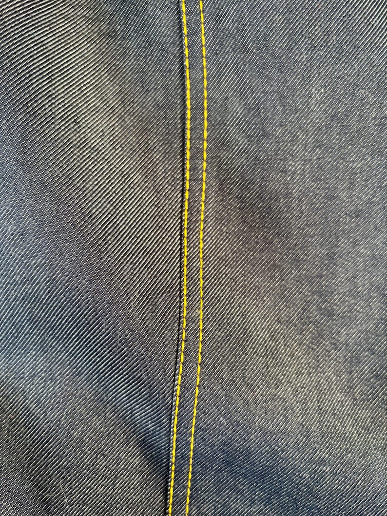 May include: Close-up of dark blue denim fabric with a yellow stitched seam running vertically. The denim has a textured appearance, with diagonal lines creating a subtle pattern. The yellow stitching provides a contrasting accent.