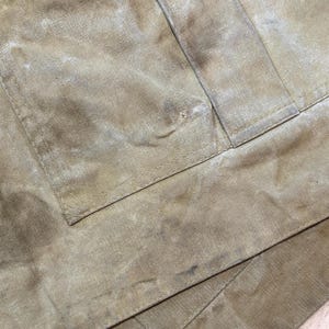 Pu&ograve; includere: Primo piano di un tessuto beige usurato, con macchie visibili e un dettaglio di tasca. Il tessuto sembra essere un materiale resistente, forse tela o un tessuto durevole simile. L'immagine evidenzia la consistenza e l'usura del materiale.