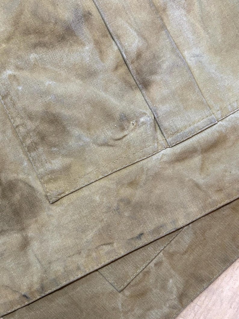 Pu&ograve; includere: Primo piano di un tessuto beige usurato, con macchie visibili e un dettaglio di tasca. Il tessuto sembra essere un materiale resistente, forse tela o un tessuto durevole simile. L'immagine evidenzia la consistenza e l'usura del materiale.