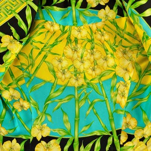 May include: Close-up of a fabric with a vibrant floral design. The pattern features yellow orchids, green bamboo stalks, and leaves against a backdrop of yellow, turquoise, and black. The edges are bordered with a geometric pattern in yellow and black.