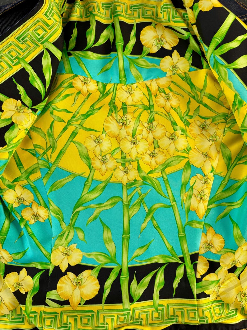 May include: Close-up of a fabric with a vibrant floral design. The pattern features yellow orchids, green bamboo stalks, and leaves against a backdrop of yellow, turquoise, and black. The edges are bordered with a geometric pattern in yellow and black.
