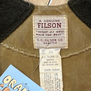 Pu&ograve; includere: Primo piano di un'etichetta di giacca Filson beige con il testo "A Genuine Filson" e "Might as well have the best". L'etichetta include anche "C.C. Filson Co. Seattle" e "Made in U.S.A." Un'etichetta con "Orange Crush" &egrave; visibile.